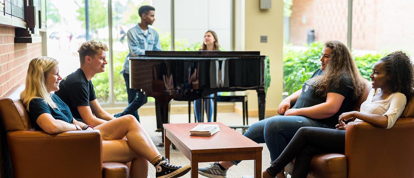 Two students at a piano while four students gather in a group in front of them.